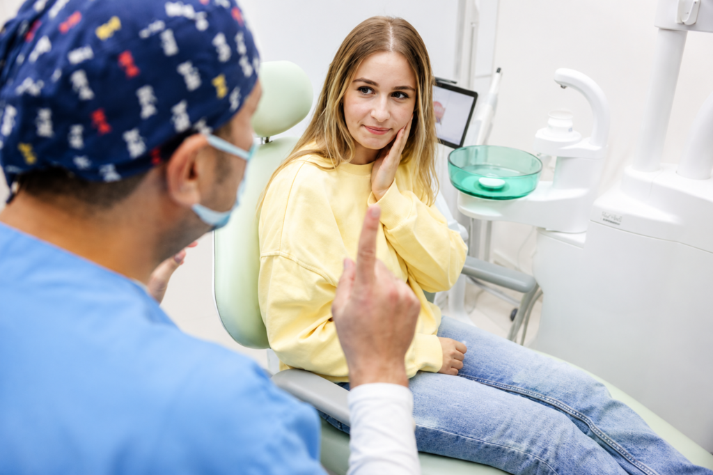Female patient holding her cheek in discomfort while consulting with a dentist in scrubs and a cap during a dental appointment