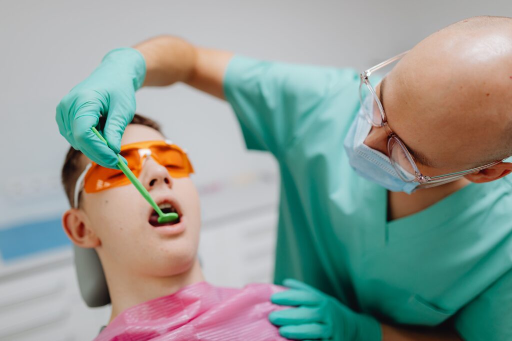 Dentist in green scrubs and protective glasses examining a young patient's teeth with a dental mirror while the patient wears orange UV safety glasses