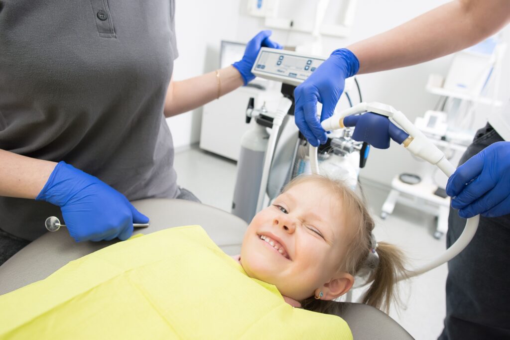 Smiling young girl in a dental chair receiving treatment from two dental staff wearing blue gloves